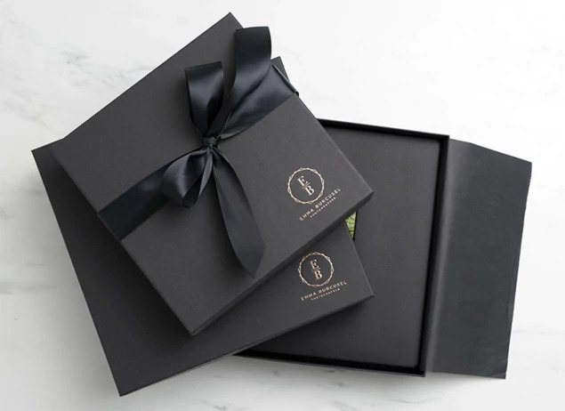Two matte black album boxes with custom foil studio logo and black satin ribbon on marble surface     Tyndell