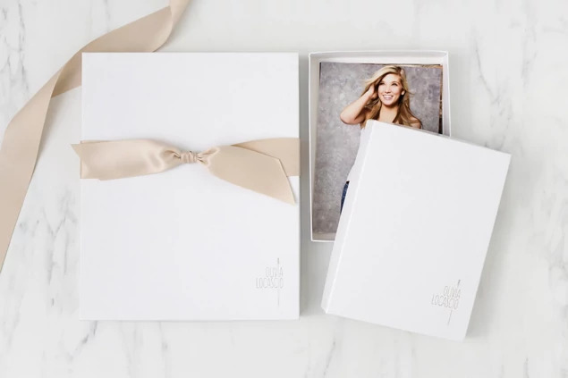 Open white leather portrait box with senior portrait print and champagne ribbon on marble surface     Tyndell