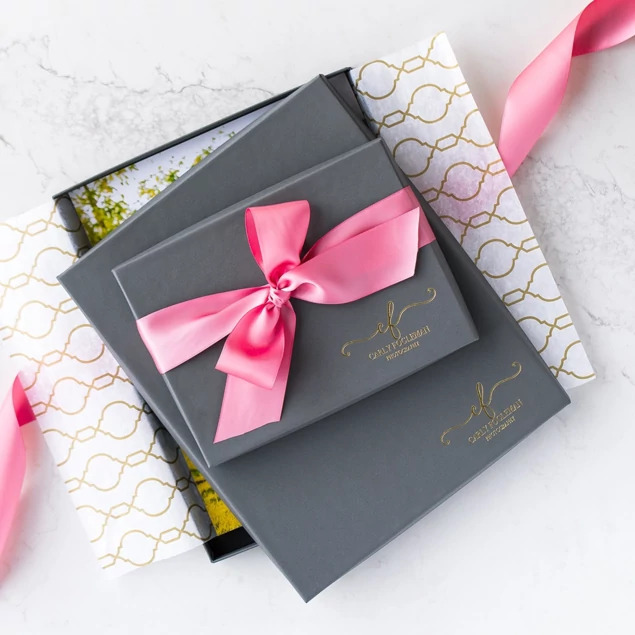 Two slate deluxe portrait boxes with custom gold foil studio logo and pink ribbon on patterned tissue paper     Tyndell