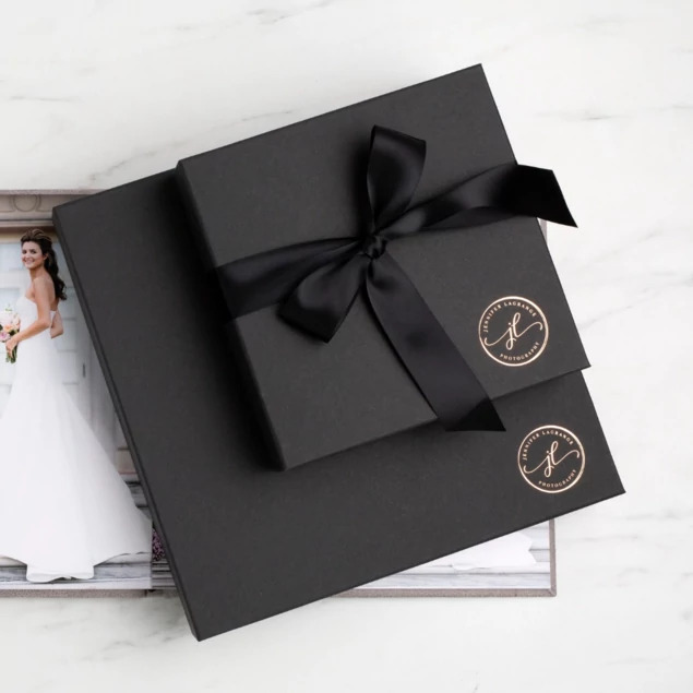 Two matte black square presentation boxes with custom foil studio logo and black satin ribbon on marble surface     Tyndell