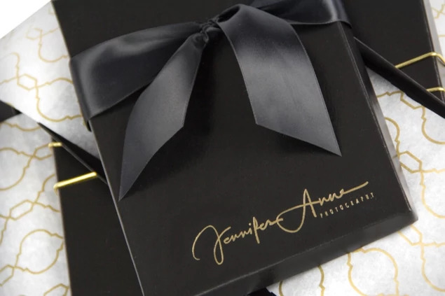 Close-up of black leather deluxe portrait box with custom gold foil studio logo and black satin ribbon     Tyndell