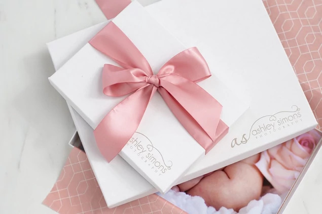Matte white deluxe portrait box with pink satin ribbon bow held above newborn baby     Tyndell newborn photography packaging