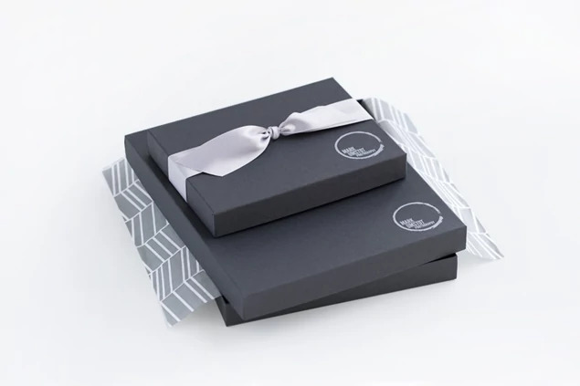 Two stacked matte black portrait boxes with custom foil studio logo and white ribbon on patterned tissue paper     Tyndell