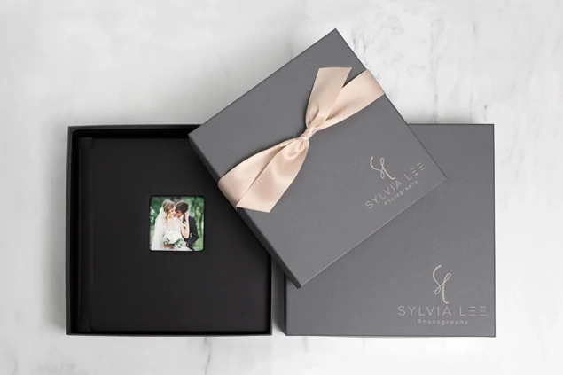 Open slate album box with wedding album inside, custom foil studio logo and champagne ribbon on marble surface     Tyndell